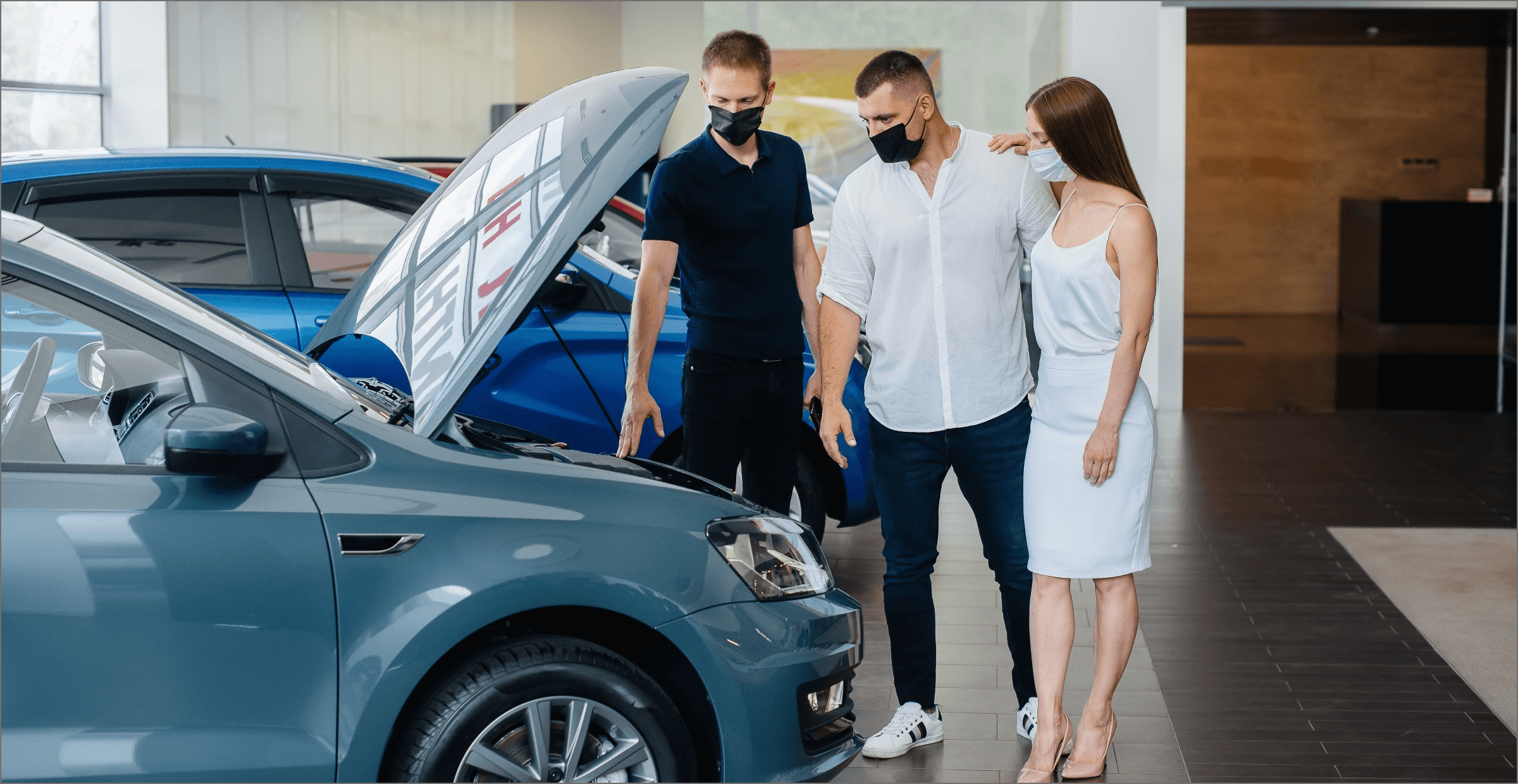 A photograph shows two men and a woman standing by a car. The car’s hood is open, and all three people are looking inside.