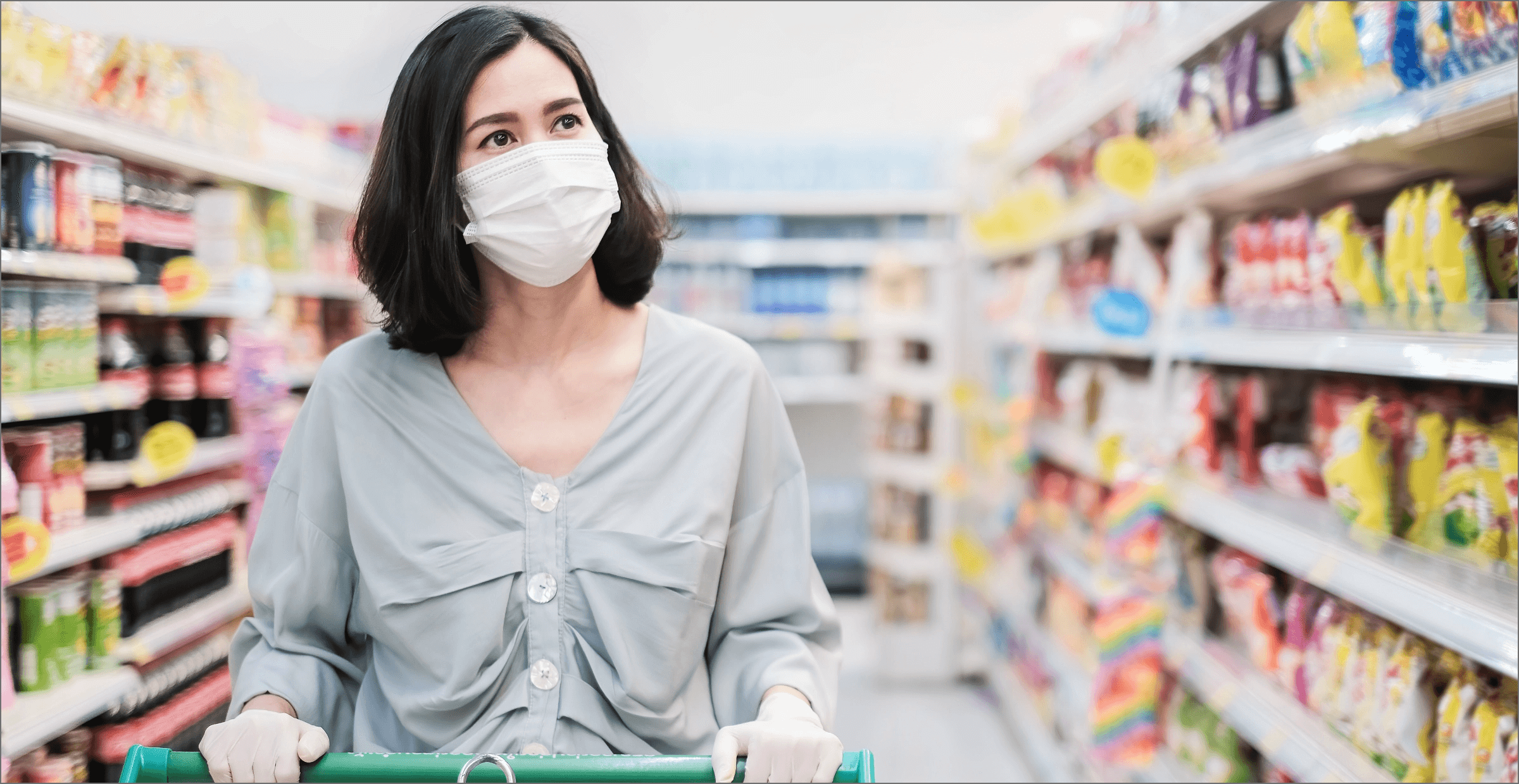 A photograph shows a woman shopping in a grocery store. She is pushing a cart, and wearing a medical facemask.